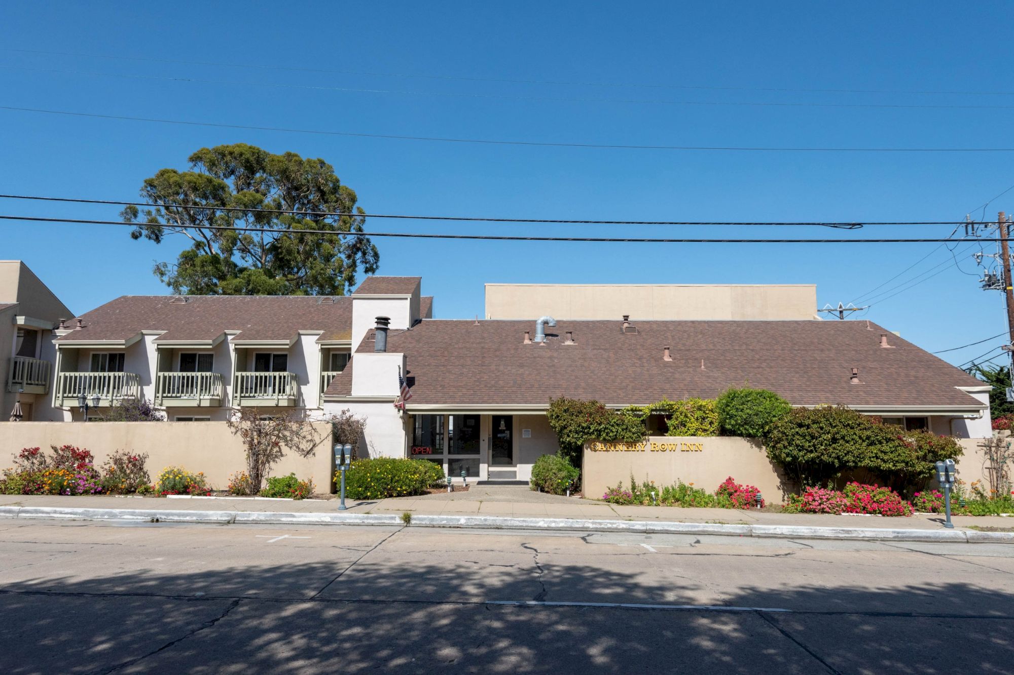 A single-story building with a sloped roof, beige walls, and a small landscaped front yard; blue sky, power lines, and a tree in the background.