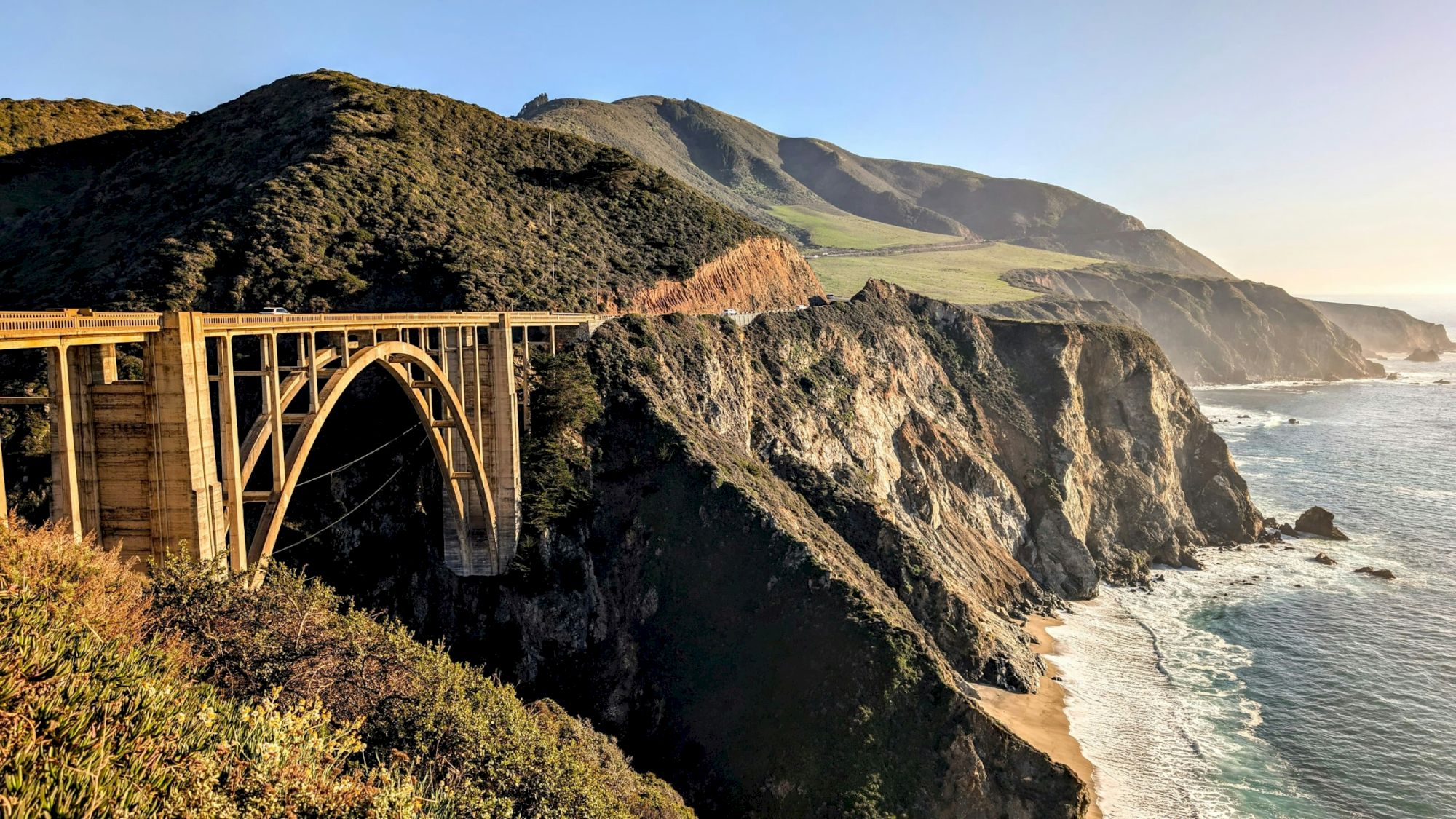 A scenic coastal view with a historic arched bridge spanning rugged cliffs, rocky coastline, and waves crashing along the shore at sunset.