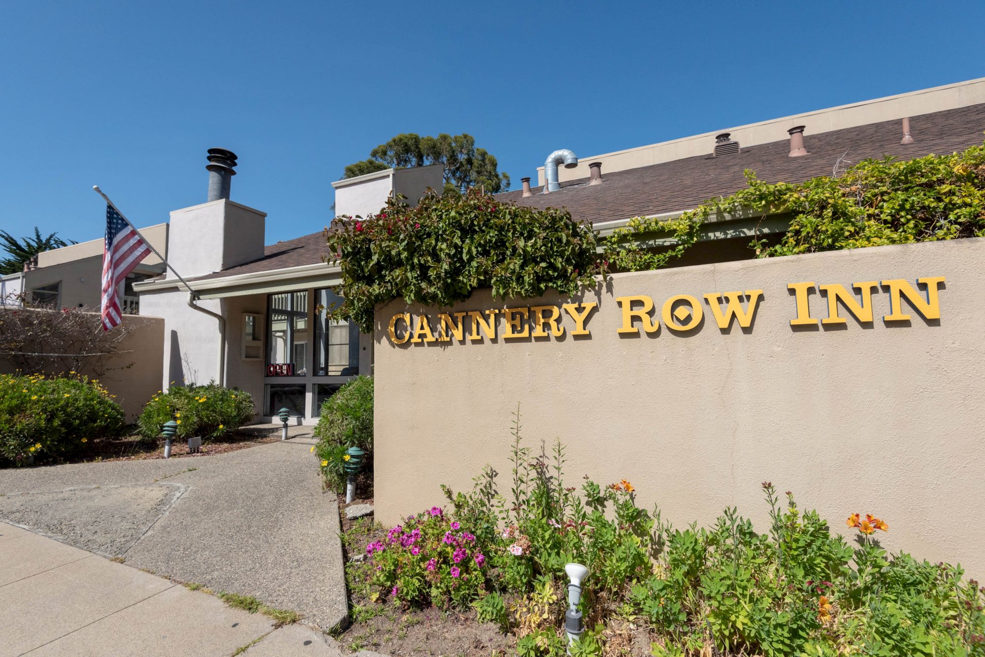 A motel entrance with a beige sign reading “CANTERY ROW INN” (likely "Cantlery"?) and a small garden in front; American flag on a pole by the doorway, clear blue sky.