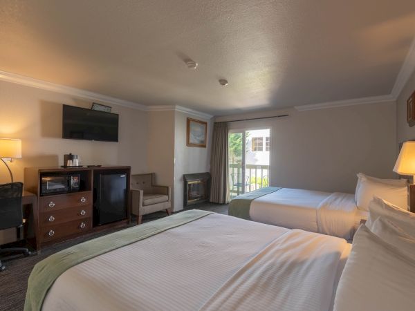 A hotel room with two beds, a desk, TV, dresser, a chair, lamps, and a small balcony outside, neatly arranged and softly lit.