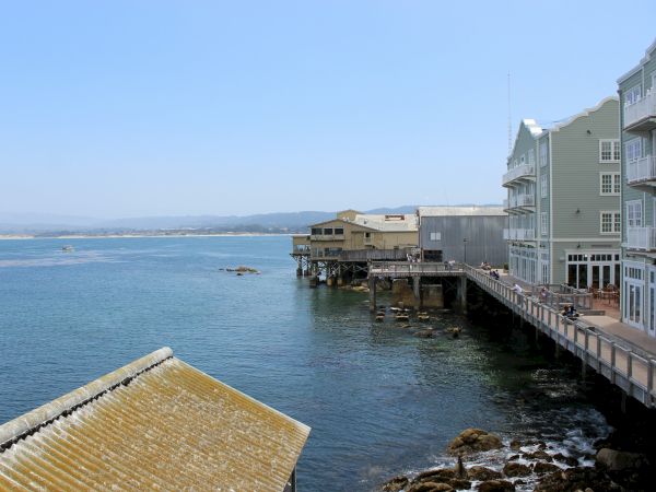 Coastal scene with a wooden pier and pastel seaside buildings over calm, blue-green water; distant hills, clear sky, rocky shoreline, tranquil ambiance.