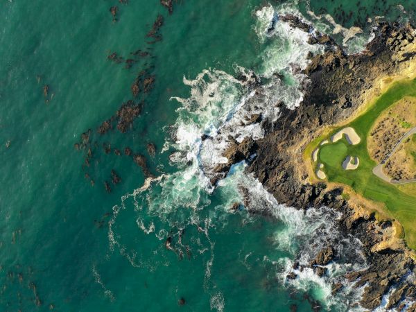 Ocean waves crash along a rocky coastline next to a grassy, sunlit cliff, with turquoise water on the left and rugged shore on the right.