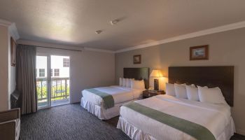 A hotel room with two large beds, side tables, lamps, a balcony door, and neutral decor with green bed runners, all under soft lighting.