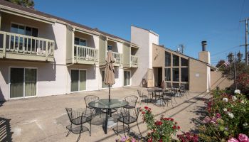 An outdoor courtyard of a two-story motel with balconies, glass doors, patio tables, chairs, a central umbrella, and blooming flowers.