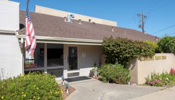 A small, single-story building with a beige facade, front porch, and an American flag; there are shrubs and a paved walkway outside, sunny day.