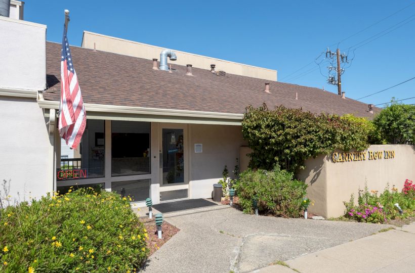 A small, single-story building with a beige facade, front porch, and an American flag; there are shrubs and a paved walkway outside, sunny day.