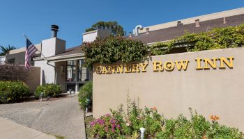 A small roadside motel named “Canterbury Row Inn” with a beige sign, greenery, and an American flag by the entrance under a clear blue sky.