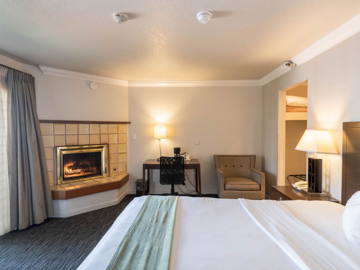 A cozy hotel room with a bed in the foreground, a fireplace to the left, a desk and chair in the back, and a small seating area with lamps by the right wall.
