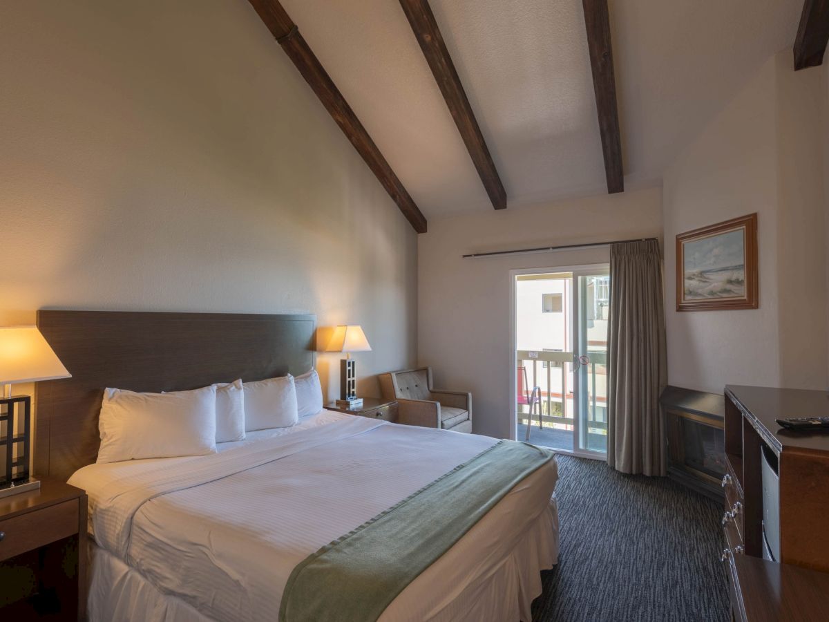 A cozy hotel room with two large beds, wooden beams, bedside lamps, a small sitting area, and a balcony door letting in natural light.