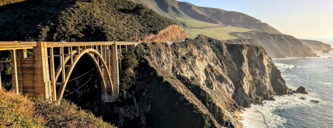A scenic bridge arches over a deep canyon with rugged cliffs and a winding road beneath, set against a sunny, mountainous backdrop.