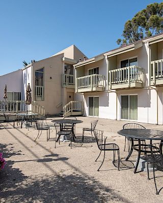A sunny courtyard outside a two-story motel or apartment complex with balconies, metal patio tables and chairs, and flowering shrubs along the edge.